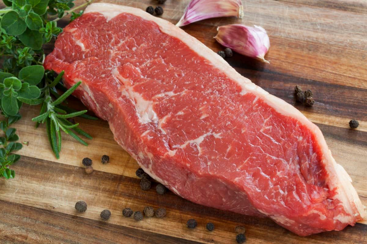 Raw sirloin steak on a wooden plank surrounded by garlic, peppercorns, and herbs